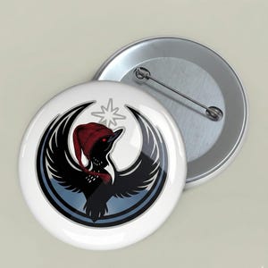 May include: A white button with a black bird design wearing a red hat and scarf, set against a crescent moon and star. The button has a silver pin back, and another button is visible in the background.