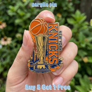 Pode incluir: Um broche de esmalte com uma bola de basquete sobre um troféu, com o texto NEW YORK KNICKS em laranja e azul. O design inclui uma silhueta do horizonte de uma cidade na base, feita em esmalte de cor dourada metálica, laranja e azul escuro.