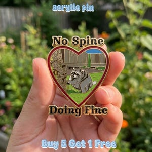 May include: A heart-shaped acrylic pin featuring a raccoon standing on green grass next to a tree trunk. The pin has a red border and gold metal edges. The text 'No Spine' is at the top and 'Doing Fine' is at the bottom in black lettering.