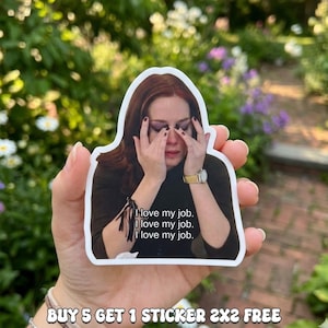 May include: A white sticker featuring a woman with red hair, covering her face with her hands. The sticker has the text "I love my job" repeated three times. The sticker is held in a hand against a blurred green background.