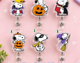 May include: Six clear acrylic badge reels featuring Snoopy in various Halloween costumes, including a vampire, a pumpkin, and a witch. Each reel has a metal clip and a retractable cord. The text Buy 5 Get 1 Free is printed at the bottom.