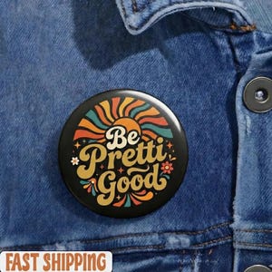 Puede incluir: Un botón negro con el texto "Be Pretti Good" en una fuente retro, rodeado de un diseño colorido de un sol, flores y espirales. El botón está sobre una chaqueta vaquera azul.