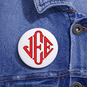 Könnte beinhalten: Ein weißer Knopf mit einem roten Monogramm-Design mit den Buchstaben J, F und E. Der Knopf ist rund und befindet sich auf einer blauen Jeansjacke.
