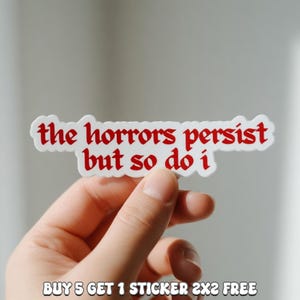 May include: A white sticker with red text that reads "the horrors persist but so do i". The sticker is held in a hand. The sticker is approximately 2x2 inches.