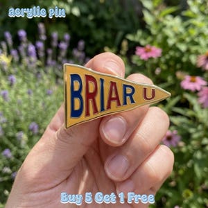 Pode incluir: Um alfinete acrílico dourado em forma de flâmula, com a palavra "BRIAR U" em letras vermelhas e azuis. O alfinete é segurado em uma mão contra um fundo de folhagem verde e flores. O texto "acrylic pin" e "Buy 5 Get 1 Free" também são visíveis.