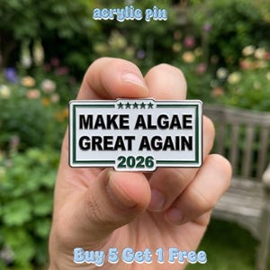 Könnte beinhalten: Ein rechteckiger Acryl-Pin mit weißem Hintergrund und dunkelgrünem Rand. Der Text lautet MAKE ALGAE GREAT AGAIN 2026 in schwarzen Großbuchstaben. Fünf kleine grüne Sterne sind über dem Text innerhalb des rechteckigen Rahmens zentriert.