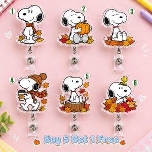 May include: Six acrylic badge reels featuring Snoopy in various autumn scenes with fall leaves, pumpkins, and coffee. Each reel is numbered one through six. The bottom text reads Buy 5 Get 1 Free. The designs include Snoopy wearing a scarf, holding a pumpkin, sitting in leaves, wearing a beanie, sitting on a tree stump, and with Woodstock.