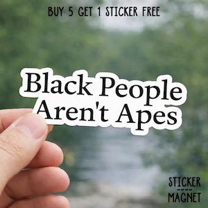 May include: A white sticker with the text "Black People Aren't Apes" in a bold, black font. The sticker is held by a hand against a blurred green background. The text "BUY 5 GET 1 STICKER FREE" is at the top.