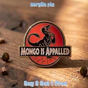 May include: A circular acrylic pin featuring a black silhouette of a dinosaur on a red background. A horizontal black banner across the center displays the white text "MONGO IS APPALED" in a bold, sans-serif font with a thin gold border.