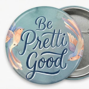 May include: A teal button with the words "Be Pretti Good" in white script and two birds with orange, purple, and blue feathers. The back of the button is silver. A decorative item.