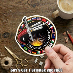 May include: A circular sticker featuring a space shuttle design with the text "PROJECT HAIL MARY." The sticker is surrounded by flags of various countries. The sticker is held by a hand. The text "BUY 5 GET 1 STICKER 2X2 FREE" is at the bottom.