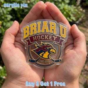 May include: Clear acrylic pin with the text "Briar U Hockey" in yellow and maroon. The pin features a hockey-themed design with a bird and crossed hockey sticks. The text "Est. 2015" is also visible.