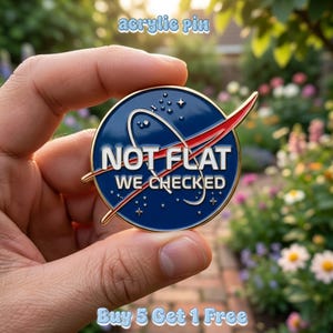 May include: A round, blue acrylic pin with a gold border, featuring the text "NOT FLAT WE CHECKED" in white. The pin has a red and white curved design, resembling a NASA logo. The words "acrylic pin" and "Buy 5 Get 1 Free" are also visible.