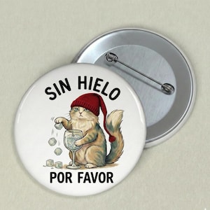 Könnte beinhalten: Ein weißer Ansteckbutton mit einer Cartoon-Katze, die eine rote Mütze trägt und ein Glas mit Eiswürfeln hält. Der Text "SIN HIELO POR FAVOR" ist in Schwarz gedruckt. Ein silberner Nadelrücken ist sichtbar.