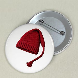 May include: A red knit hat with a long, braided tail and tassel, displayed on a white button. The hat is a vibrant shade of red, with a ribbed brim and a pointed crown. The button is round and has a metal pin on the back.