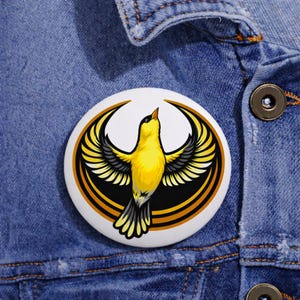Pode incluir: Um botão redondo branco com um pássaro amarelo com asas pretas e brancas, em um design circular preto e dourado. O pássaro é retratado com as asas abertas, em uma jaqueta jeans.