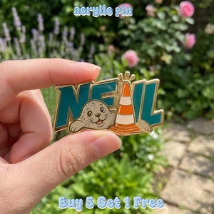 May include: An acrylic pin featuring the word NEAL in teal block letters with a gold outline. A cartoon seal with a smiling face is positioned in front of the letters, next to an orange and white traffic cone. The pin has a gold metal finish.