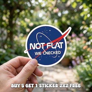 May include: A round sticker with a blue background and the NASA logo, featuring the text "NOT FLAT WE CHECKED" in white. The sticker has a red swoosh design and white stars. The sticker is held in a hand against a blurred outdoor background.