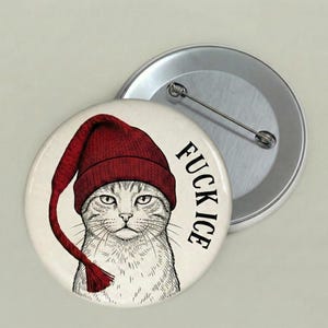 Może przedstawiać: Okrągła przypinka z ilustracją kota w czerwonej dzianinowej czapce. Na przypince widnieje czarny napis "FUCK ICE". W tle widoczna jest druga, srebrna przypinka.