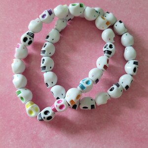 Könnte beinhalten: Ein weißes Perlenarmband mit schädelartigen Perlen. Die Schädel haben schwarze Augen und sind mit verschiedenen Farben verziert, darunter Rosa, Blau, Grün und Gelb. Das Armband wird vor einem rosa Hintergrund gezeigt.