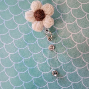 May include: A white and brown crocheted flower badge reel with a clear plastic retractable cord and a metal clip. The flower has a brown centre and white petals. The background is a teal, scale-patterned surface.