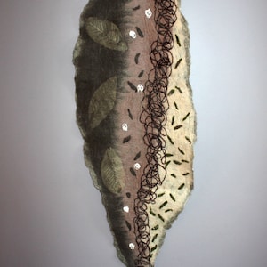 May include: A brown and green felt wall hanging with a twig at the top. The hanging has a vertical design with leaves, white dots, and brown string. The hanging is on a light purple wall.