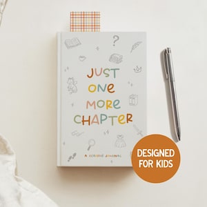 May include: A white reading journal with the text "JUST ONE MORE CHAPTER" in colourful letters. The journal has small illustrations and a bookmark. A silver pen rests beside the journal. A round orange sticker says "DESIGNED FOR KIDS".