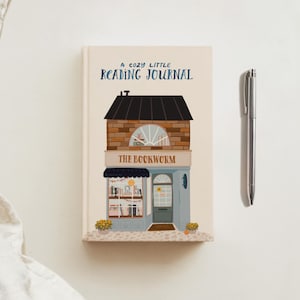 May include: A reading journal with a bookstore illustration. The cover reads "A Cozy Little Reading Journal" and "The Bookworm". A silver pen lies next to the journal on a white surface. The journal is for recording reading notes.