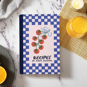 Peut inclure: Un livre de recettes avec une bordure à carreaux bleus et blancs et un centre rose clair. La couverture présente une illustration de tomates et le texte "RECIPES by MARIA GHIEPPE". Un verre de jus d'orange, une bougie et des tranches d'orange sont également visibles.