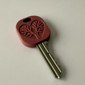May include: A red key with a heart-shaped design featuring a tree. The key's head is a solid red color, and the metal blade extends from the bottom. The tree design is embossed within the heart shape.