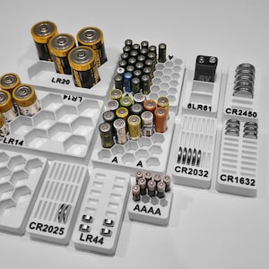 May include: A collection of white battery organizers filled with various battery types, including AA, AAA, and others. The organizers are labeled with battery sizes like LR20, LR14, CR2025, and 18650. The batteries are gold and black.
