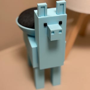 May include: A light blue, block-shaped toy animal with a rounded head and black eyes. The toy has a small, black, round object on its back.