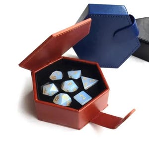 Puede incluir: Una caja de dados hexagonal roja con interior negro, abierta para revelar un juego de dados azul claro con numeración dorada. En el fondo, dos cajas hexagonales cerradas en azul y negro. Los dados son para juegos de mesa.