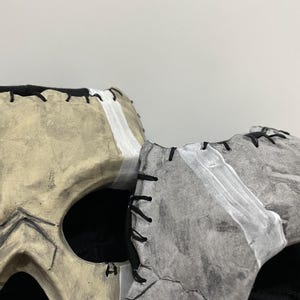 May include: Two skull masks, one beige and one grey, are displayed side-by-side. The beige mask has black stitching and white accents. The grey mask has black stitching and white accents. Both masks have large eye openings and are set against a plain background.