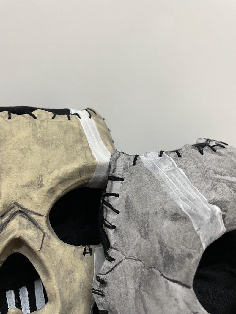 May include: Two skull masks, one beige and one grey, are displayed side-by-side. The beige mask has black stitching and white accents. The grey mask has black stitching and white accents. Both masks have large eye openings and are set against a plain background.