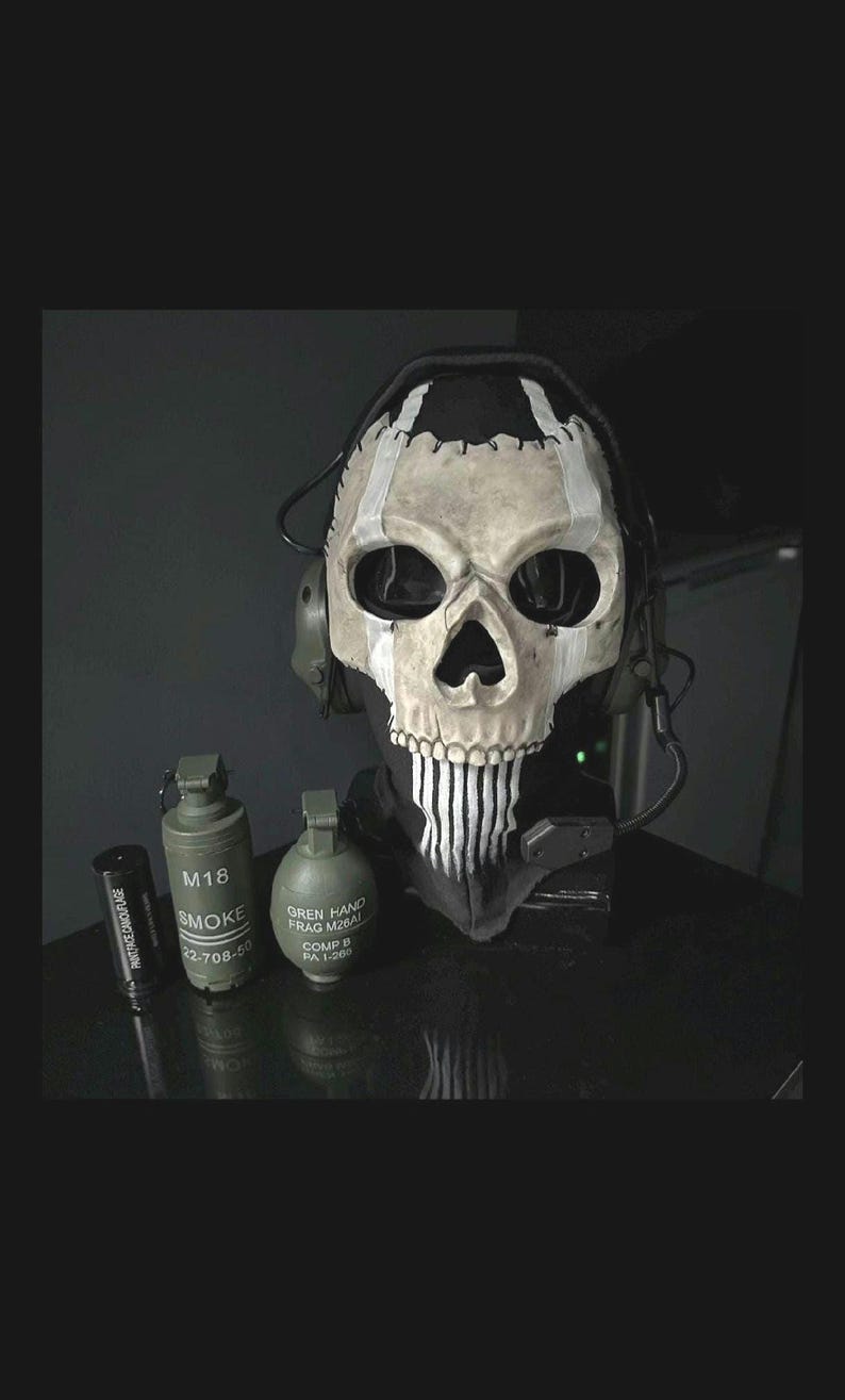 May include: A skull mask with a black neck covering and attached headset. The mask is white with black accents and a stitched design. Two green grenades and a black cylinder are in front of the mask.