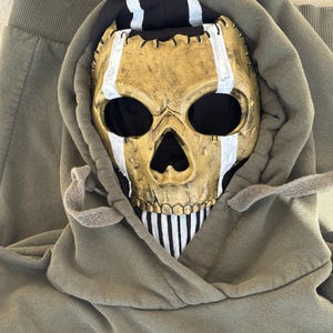 May include: A gold skull mask with white stripes and black eye sockets sits inside the hood of a green hoodie. The hoodie has drawstrings and the letters "MP" printed on the front.