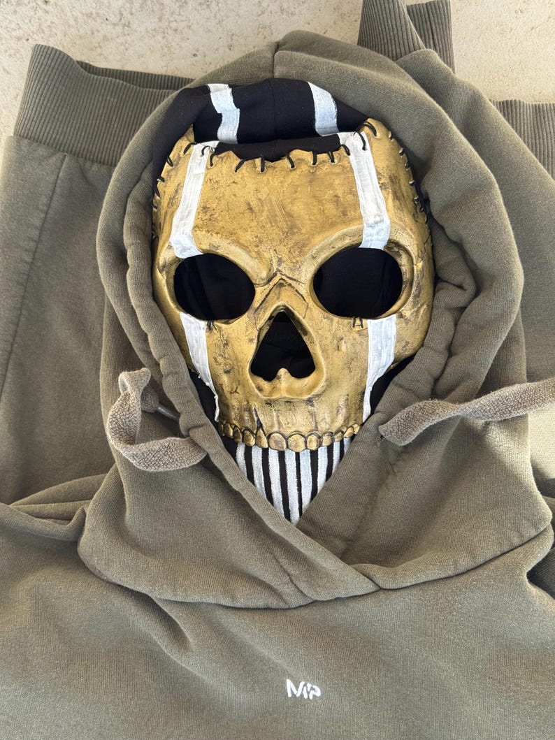 May include: A gold skull mask with white stripes and black eye sockets sits inside the hood of a green hoodie. The hoodie has drawstrings and the letters "MP" printed on the front.