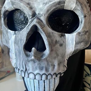 May include: A grey skull mask with black eye sockets and a black interior. White accents are painted on the skull, and the mask is stitched together with black thread. The mask is designed for Halloween or costume use.