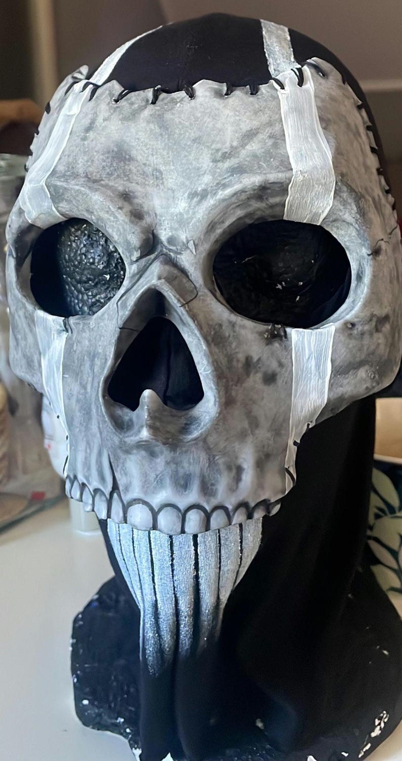 May include: A grey skull mask with black eye sockets and a black interior. White accents are painted on the skull, and the mask is stitched together with black thread. The mask is designed for Halloween or costume use.