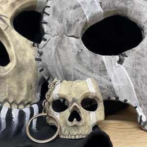 May include: A skull-shaped keyring sits on a black cloth, with two skull masks in the background. The masks and keyring are painted with white stripes and have a distressed, aged appearance. The masks have stitched edges.