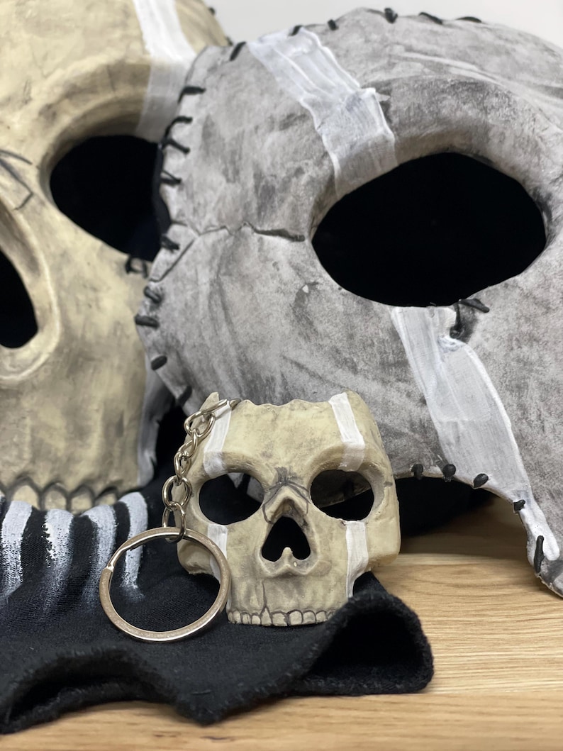 May include: A skull-shaped keyring sits on a black cloth, with two skull masks in the background. The masks and keyring are painted with white stripes and have a distressed, aged appearance. The masks have stitched edges.