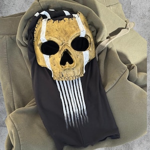 May include: A gold skull mask with white detailing and black eye sockets sits on a black top with white vertical stripes. The top is on a green hoodie and matching tracksuit bottoms.