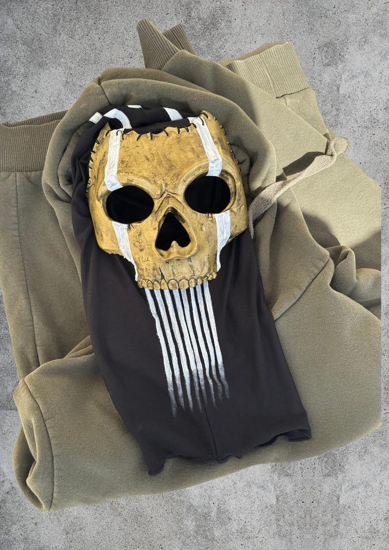 May include: A gold skull mask with white detailing and black eye sockets sits on a black top with white vertical stripes. The top is on a green hoodie and matching tracksuit bottoms.