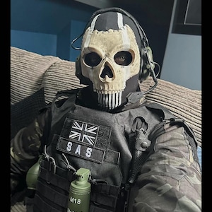 May include: A person wearing a skull mask, black tactical vest, and camouflage jacket. The mask has a skull design with black eye sockets and a white jawline. The vest has a patch that says "SAS" and a British flag. A green grenade is attached to the vest.