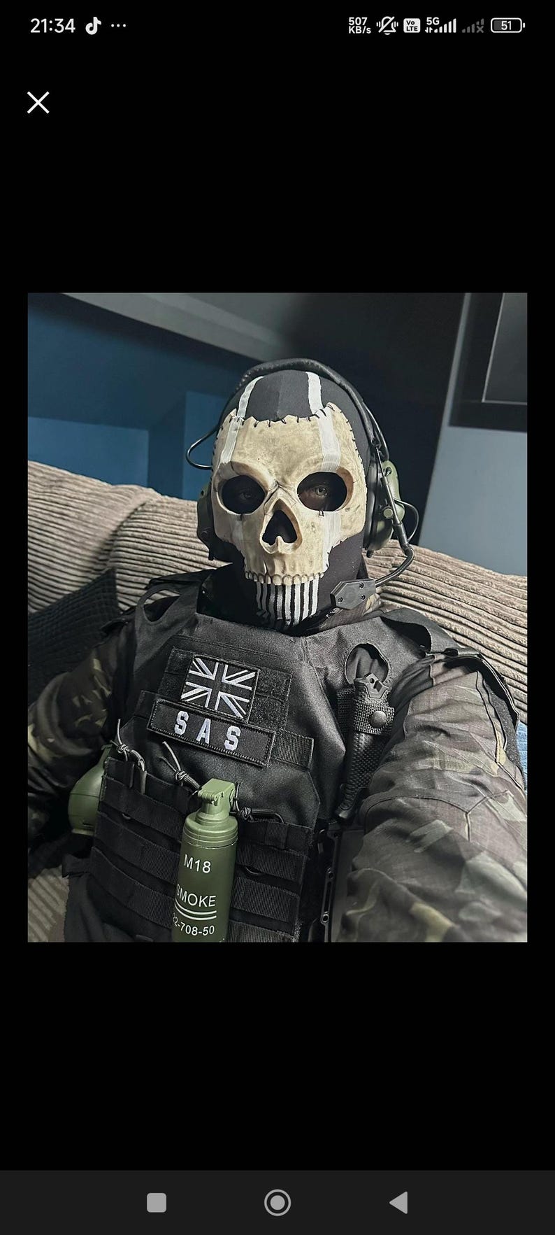 May include: A person wearing a skull mask, black tactical vest, and camouflage jacket. The mask has a skull design with black eye sockets and a white jawline. The vest has a patch that says "SAS" and a British flag. A green grenade is attached to the vest.