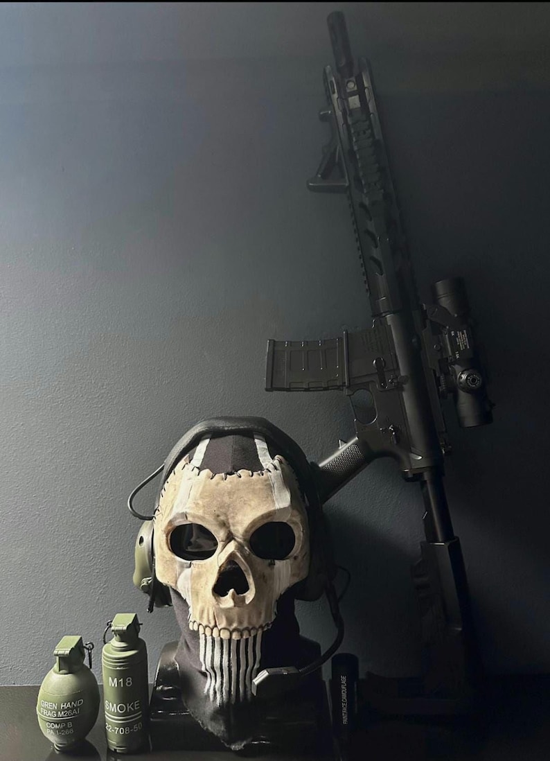 May include: A black rifle with a scope stands against a dark wall beside a skull mask with headphones. Two green grenades and a small black object are in front of the mask. The mask has a black hood and white stripes.