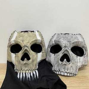 May include: Two skull masks with large eye sockets and open mouths. One mask is tan with white accents and a black fabric drape. The other mask is grey with white accents. Both masks have stitched detailing.