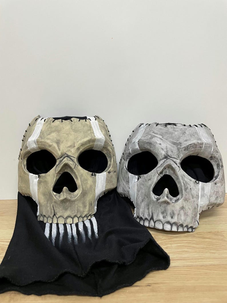 May include: Two skull masks with large eye sockets and open mouths. One mask is tan with white accents and a black fabric drape. The other mask is grey with white accents. Both masks have stitched detailing.