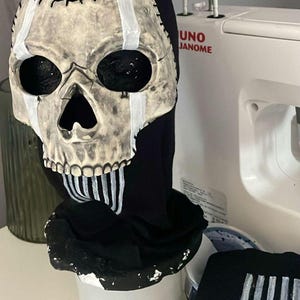 May include: A skull mask with a black hood. The skull is painted in a bone-like colour with black eye sockets and a large nose opening. White stripes accent the skull and the hood. The mask is likely for Halloween or cosplay.